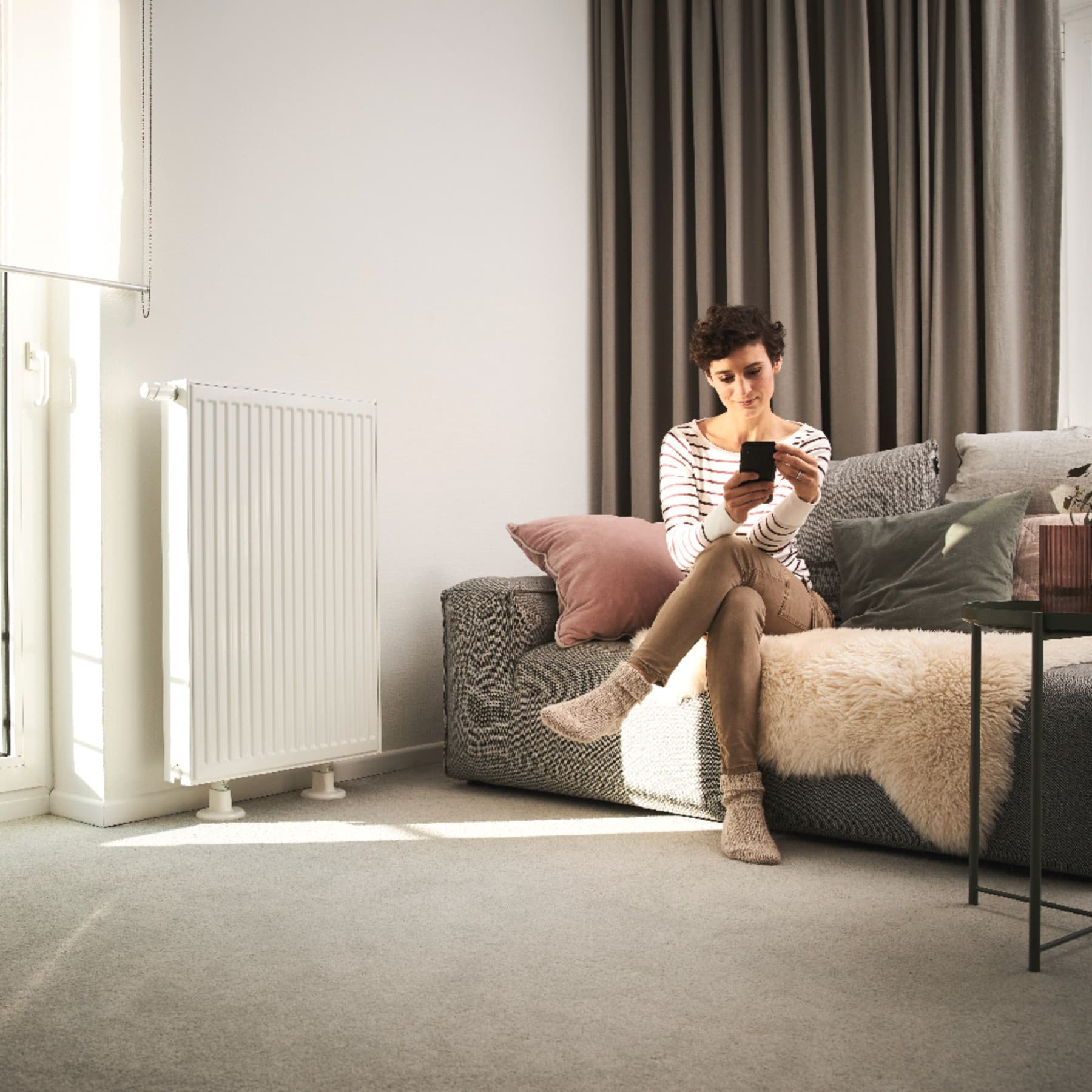 Vrouw zit op bank naast radiator en kijkt naar mobiele telefoon
