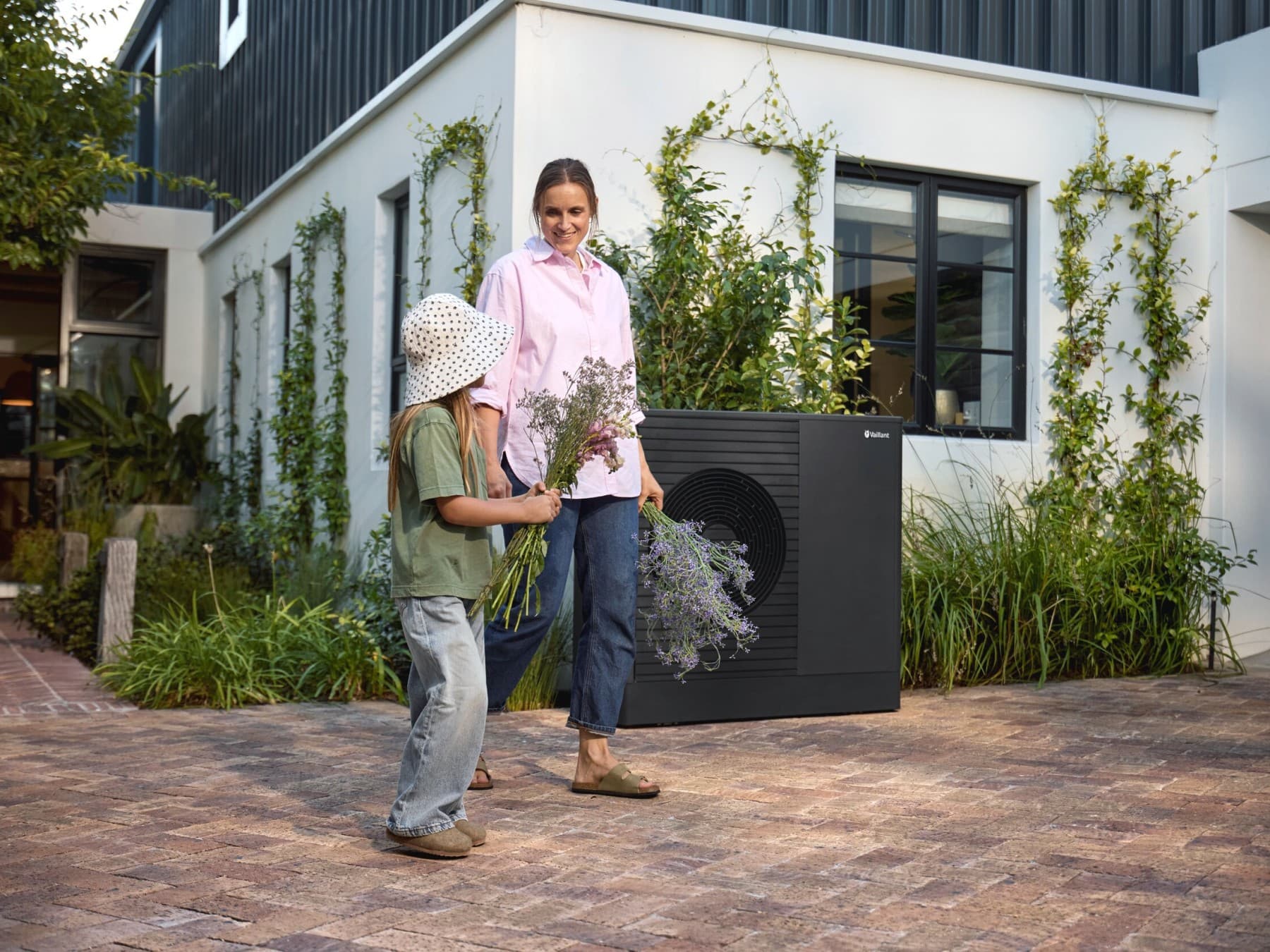 Een vrouw en een kind staan voor een huis met een warmtepomp, met bloemen in hun handen.